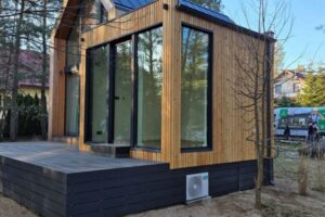 Nowoczesny domek modułowy z dużymi oknami i drewnianą elewacją, taras wypoczynkowy oraz minimalistyczna architektura harmonijnie wpisana w naturalne otoczenie lasu