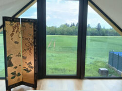 Panoramiczne okno w sypialni domu modułowego Perfect Cube w Swisttal zapewniające widok na zielone tereny i naturalne doświetlenie wnętrza.